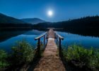 A pier leads across the surface of a mirror-smooth, moonlit lake at nighttime.
