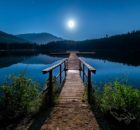 A pier leads across the surface of a mirror-smooth, moonlit lake at nighttime.