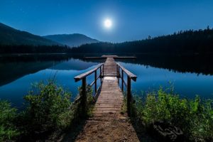 A pier leads across the surface of a mirror-smooth, moonlit lake at nighttime.